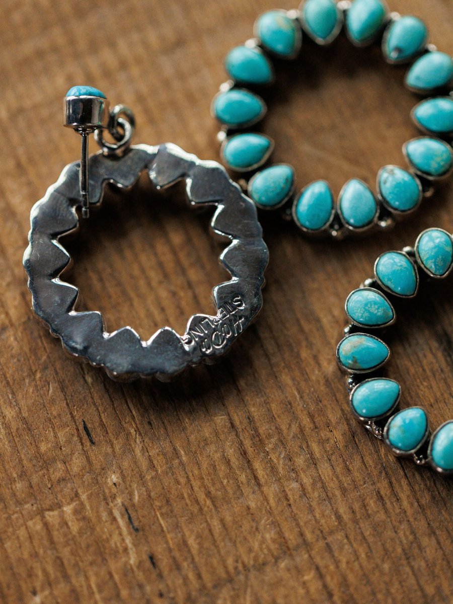 Close-up of the Ocala Earrings by Savannah Sevens western life{&}style featuring sterling silver, Kingman turquoise stones in a floral design on a wooden surface. One earring is flipped, showing a jagged back and stamped maker’s mark.