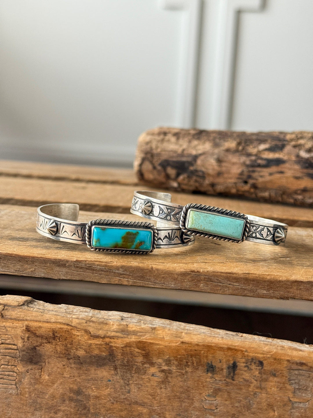 Two Ocala Turquoise Cuff bracelets by Savannah Sevens western life{&}style, crafted in sterling silver with engraved patterns and rectangular Kingman turquoise stones, rest on rustic wooden surfaces against a blurred wooden background.