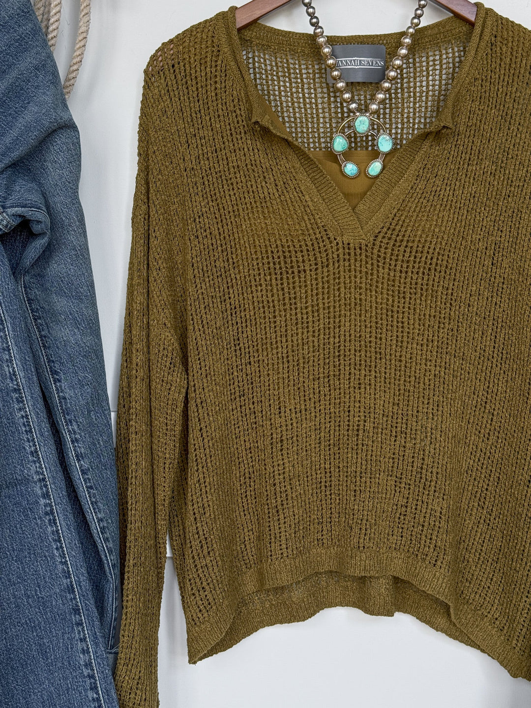 The Ocilla Olive Open Knit Top by Savannah Sevens western life{&}style, featuring long sleeves and a v-split neckline, is displayed on a hanger and styled with a turquoise beaded necklace and blue jeans beside it.