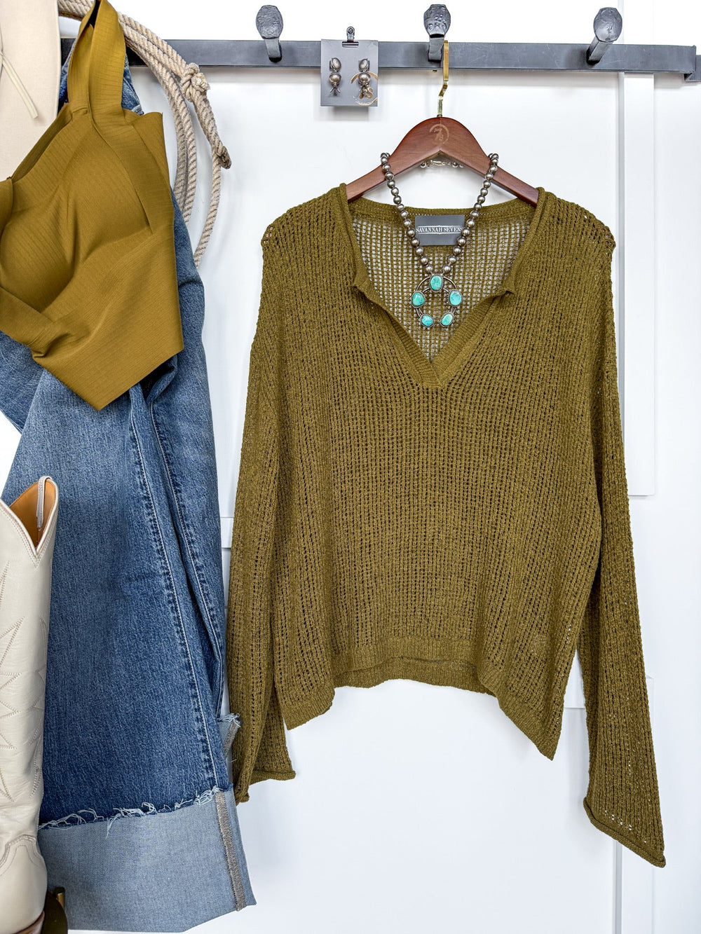 The Ocilla Olive Open Knit Top from Savannah Sevens western life{&}style, featuring a v-split neckline and long sleeves, is displayed on a hanger and accented with a turquoise necklace. Nearby, blue jeans, a matching brami top, a cream bag, and boots are neatly arranged on wall hooks.