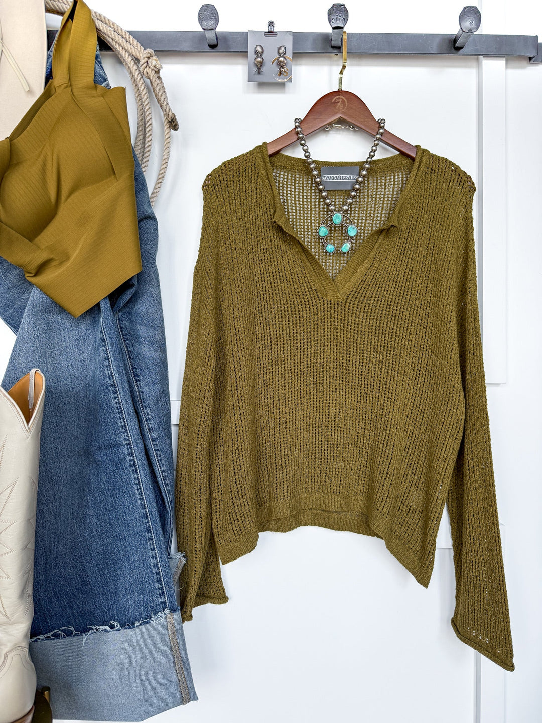 The Ocilla Olive Open Knit Top from Savannah Sevens western life{&}style, featuring a v-split neckline and long sleeves, is displayed on a hanger and accented with a turquoise necklace. Nearby, blue jeans, a matching brami top, a cream bag, and boots are neatly arranged on wall hooks.