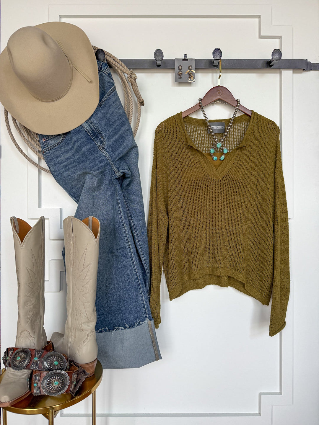 The Ocilla Olive Open Knit Top by Savannah Sevens western life{&}style, featuring long sleeves and a v-split neckline, hangs alongside tan cowboy hat and jeans on a wall hook. Below, cream cowboy boots and turquoise jewelry are arranged on a stool. The earthy tones and textures create a distinctly western, boho-chic vibe.