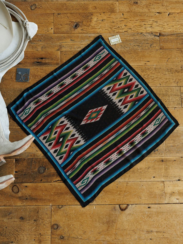 The Oconee Neck Scarf by Savannah Sevens western life{&}style features jewel-toned geometric patterns and is shown on a rustic wooden floor alongside a white hat, white boots, a tag, and silver earrings.