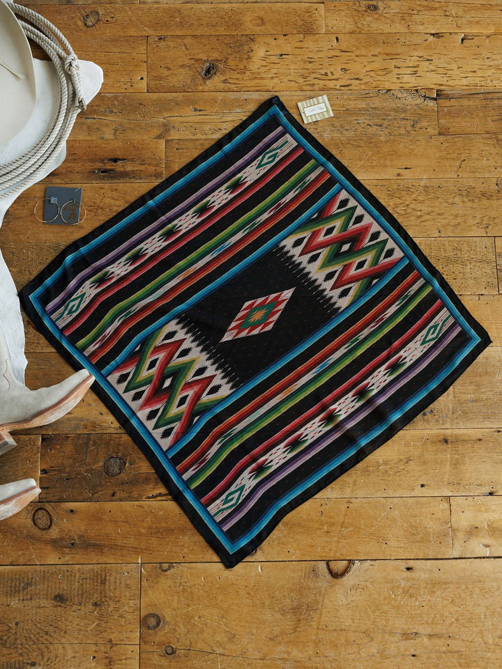The Oconee Neck Scarf by Savannah Sevens western life{&}style features jewel-toned geometric patterns and is shown on a rustic wooden floor alongside a white hat, white boots, a tag, and silver earrings.