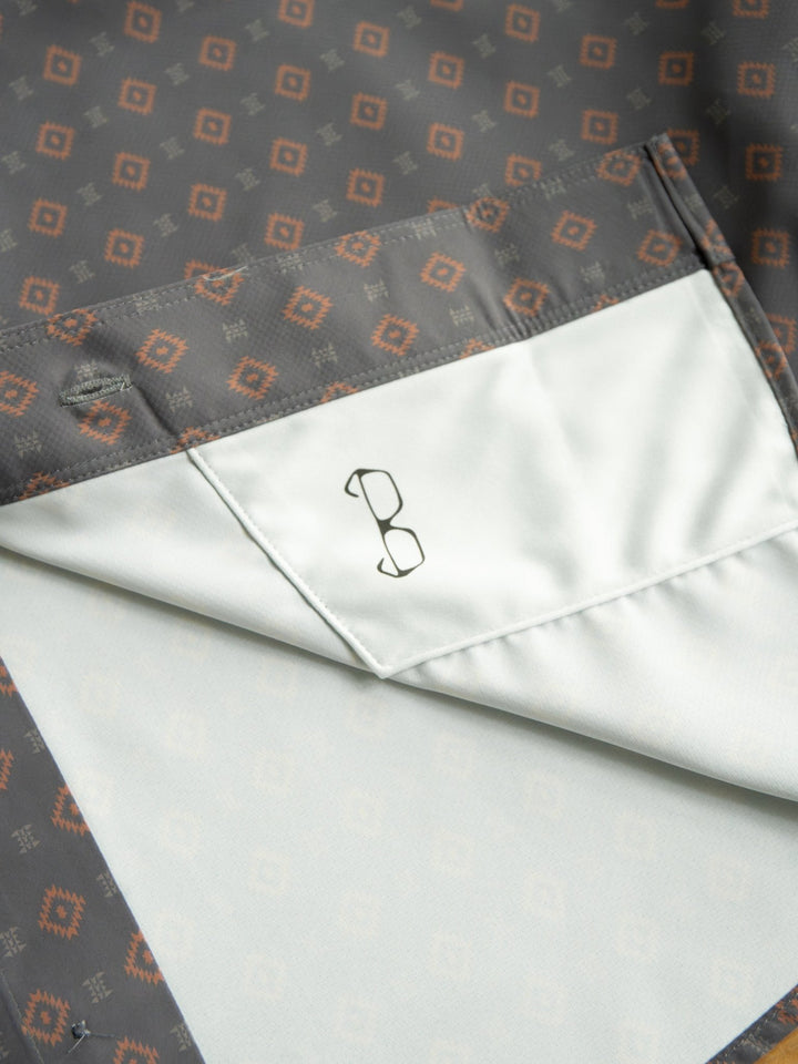 A close-up of the Odem Ariat Men's Southwest Button Up by Savannah Sevens western life{&}style, showing its patterned gray and orange fabric, white inner pocket, and a minimalist eyeglasses icon printed on the pocket.