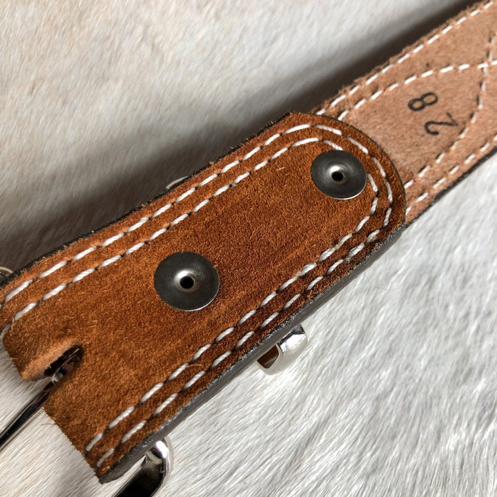 A close-up of the Okalla Belt by Savannah Sevens western life{&}style, featuring brown rough out leather, white stitching, metal rivets, and a sterling silver buckle; the number 28 is stamped on the belt.