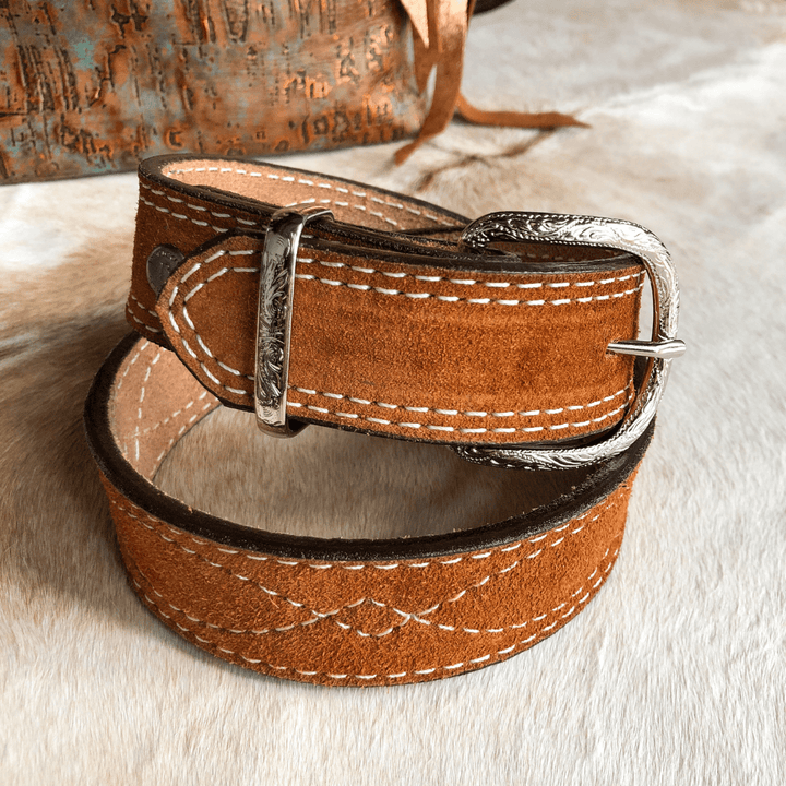 The Okalla Belt by Savannah Sevens western life{&}style, featuring brown rough out leather, white stitching, and an engraved sterling silver buckle, is coiled on a light fur surface with a rustic textured bag in the background.