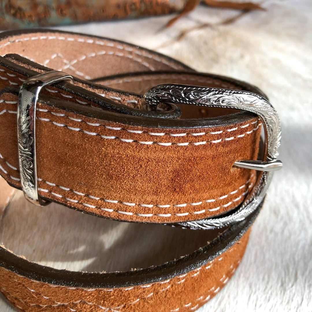 A close-up of the Okalla Belt by Savannah Sevens western life{&}style, featuring brown rough out leather, white stitching, and an ornate sterling silver engraved buckle.