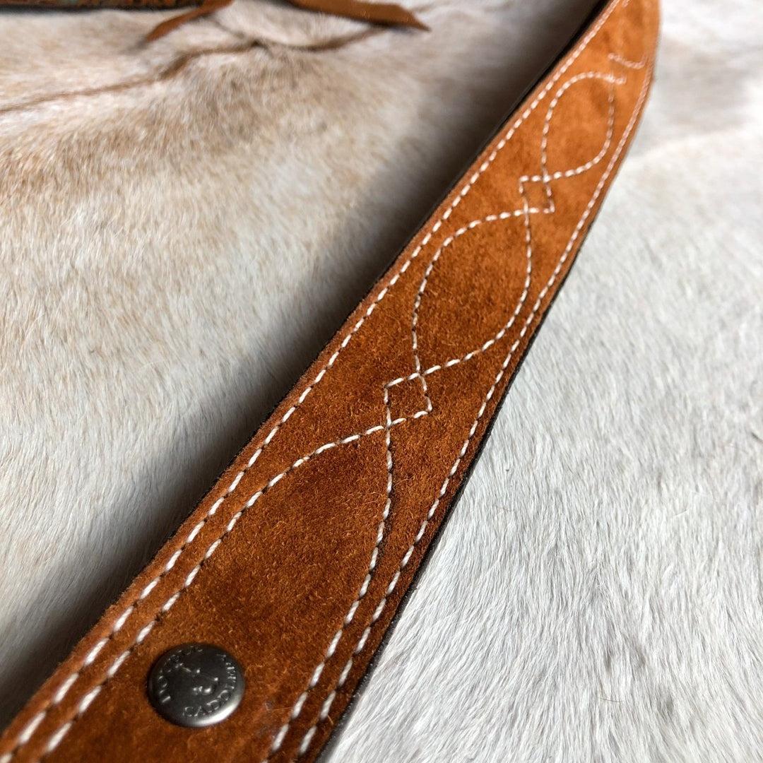 A close-up of the Okalla Belt by Savannah Sevens western life{&}style, featuring brown leather with decorative white looped stitching and a visible metal rivet, resting on a light surface.