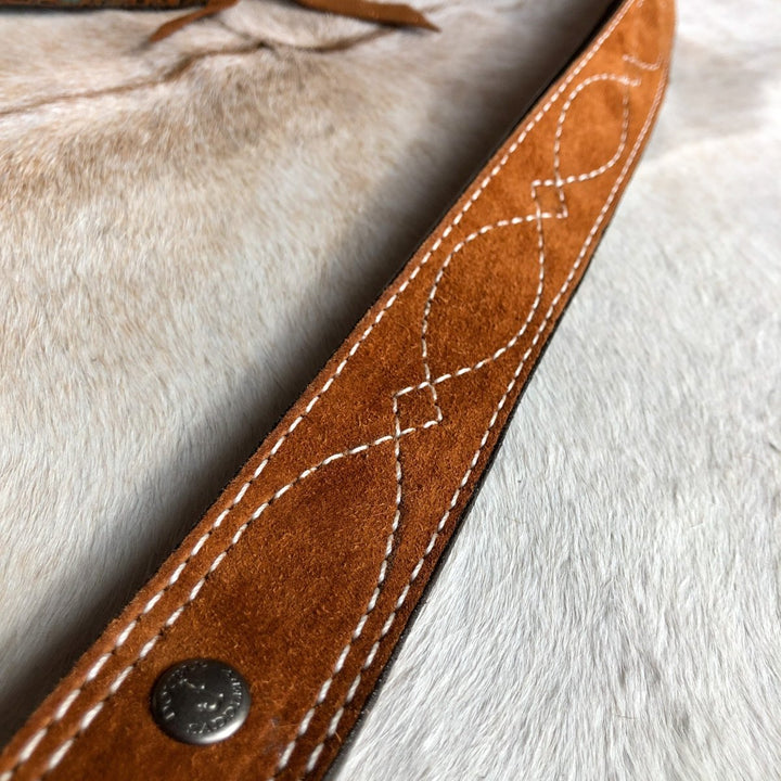 A close-up of the Okalla Belt by Savannah Sevens western life{&}style, featuring brown leather with decorative white looped stitching and a visible metal rivet, resting on a light surface.