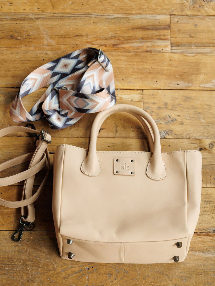 The Oketo Buckskin Leather Handbag by Savannah Sevens western life{&}style is a beige concealed carry crossbody with short handles, metal feet, and two detachable straps—one plain beige, one neutral geometric pattern—shown on a wooden surface.