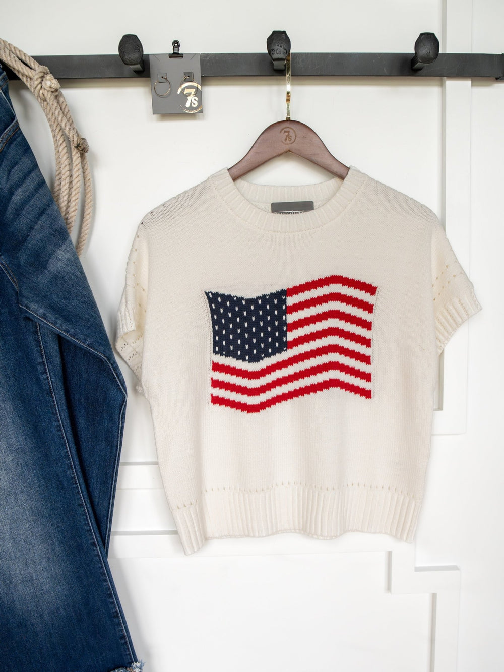 The Old Glory American Flag Knit Top by Savannah Sevens western life{&}style, a white cotton-blend short sleeve, hangs on a hanger against a white wall. A denim jacket and rope are partially visible to the left.