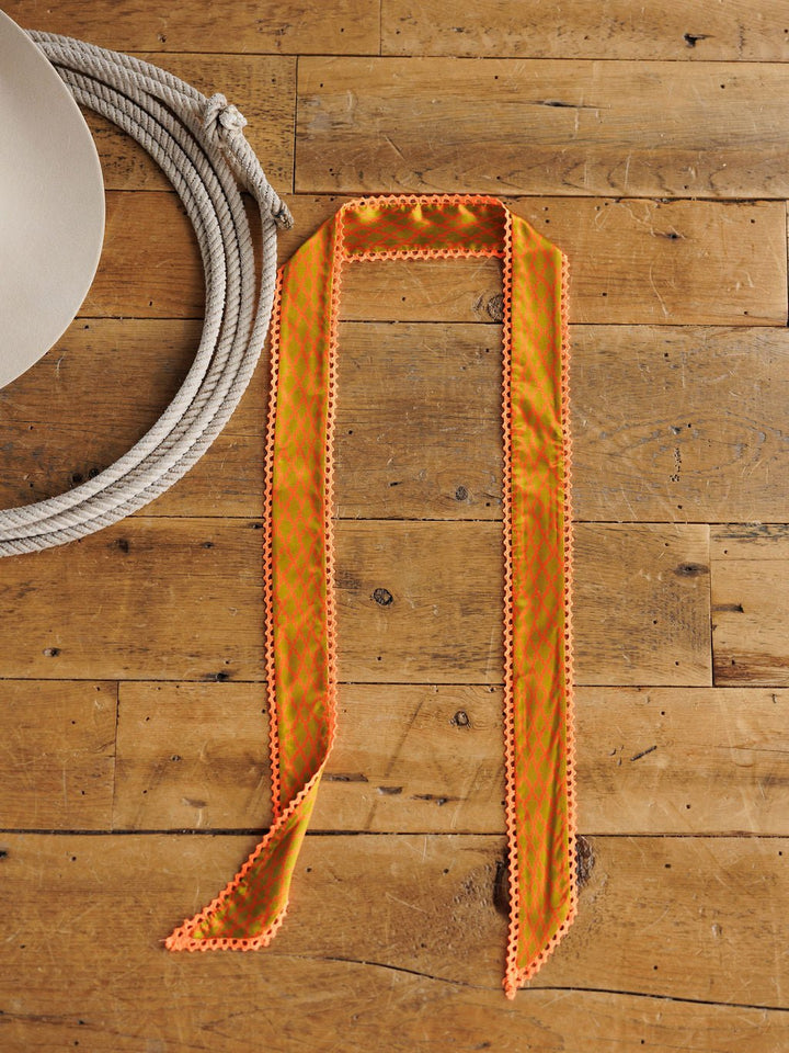 The Olive You Twilly by Savannah Sevens western life{&}style, a long satin accessory with an orange and green pattern and crochet trim, is displayed on a wooden floor beside a coiled white rope and part of a beige hat.