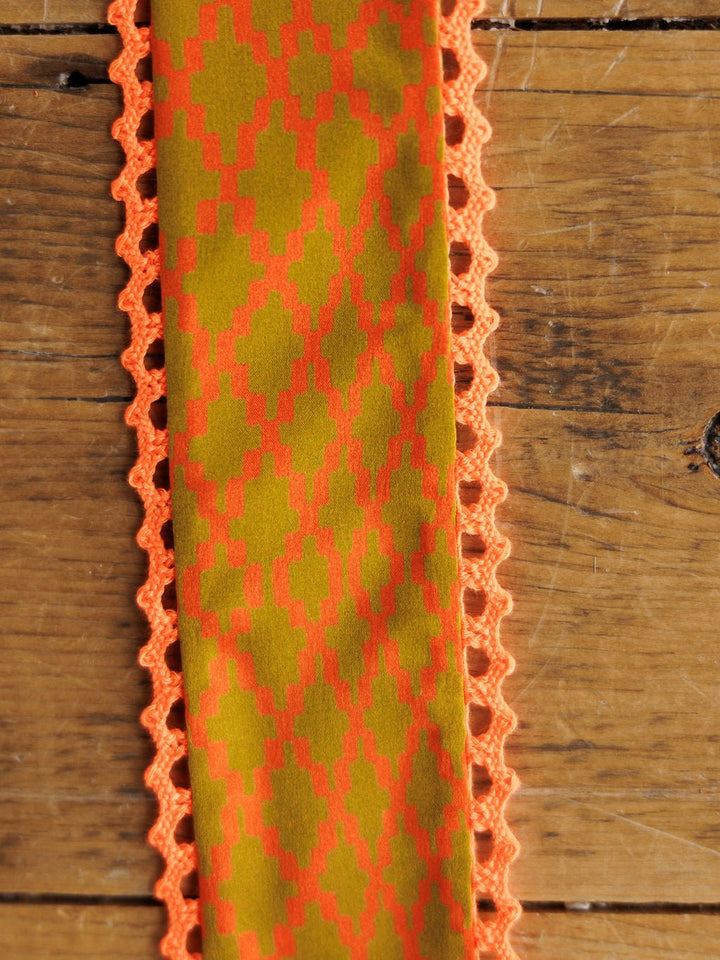The Olive You Twilly by Savannah Sevens western life{&}style features yellow-green satin with an orange geometric pattern and delicate orange lace trim on both sides, displayed on a wooden surface.