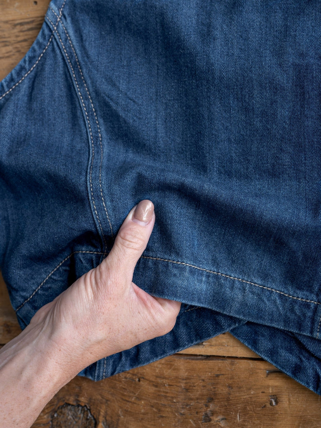 A hand pinches the hem of The Osage medium dark wash blue denim jeans by Savannah Sevens western life{&}style, shown on a wooden surface.