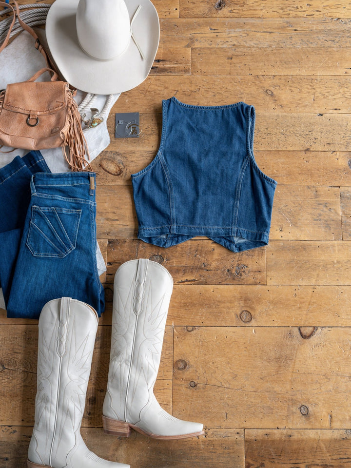 Flat lay of The Osage by Savannah Sevens western life{&}style: white cowboy hat, tan fringe purse, denim crop vest (medium dark wash), blue jeans, white cowboy boots, and silver jewelry on wooden floor.