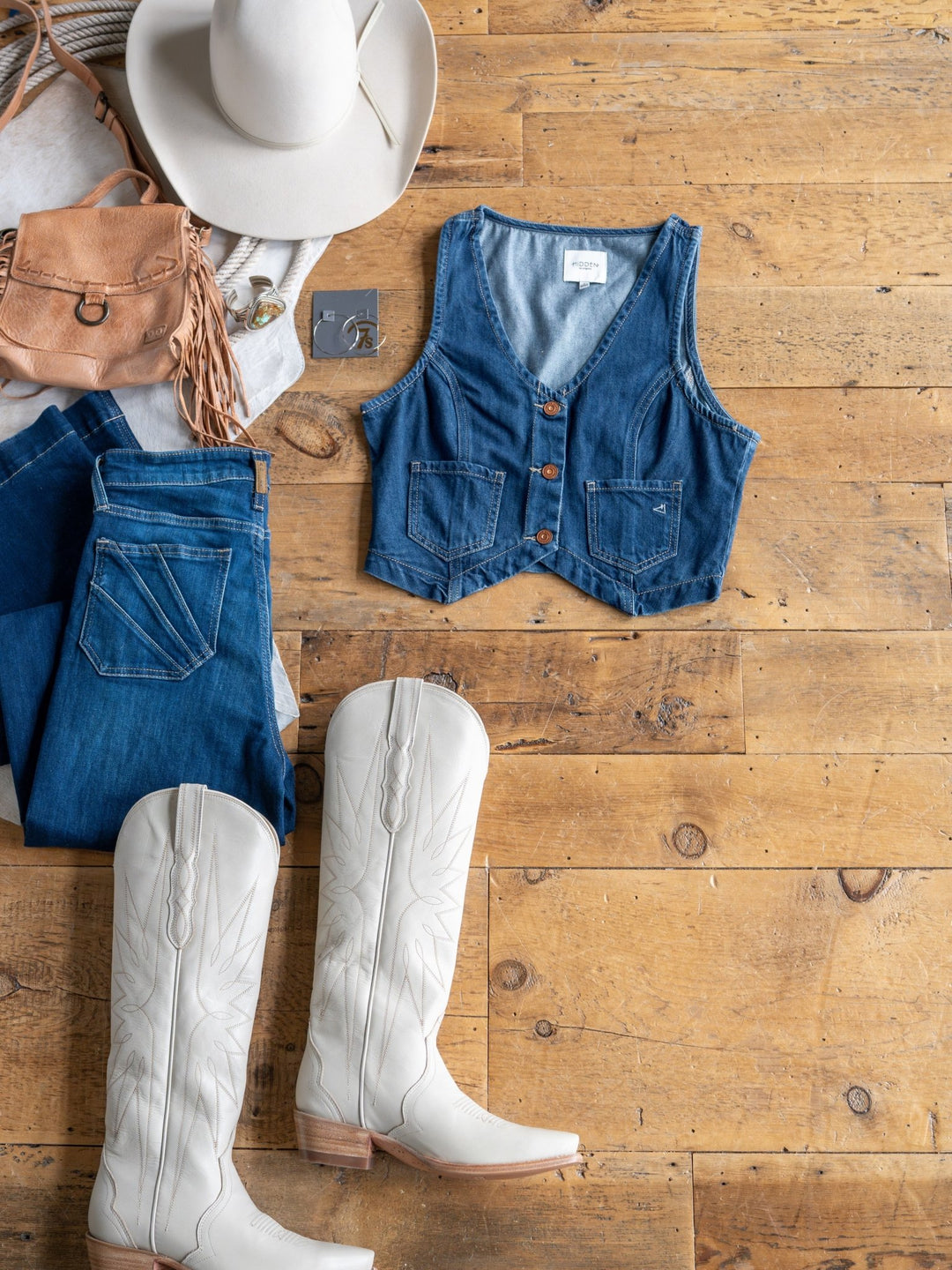 Flat lay on wooden floor featuring The Osage white cowboy boots from Savannah Sevens western life{&}style, medium dark wash jeans, a denim crop vest, white cowboy hat, tan fringe purse, and gray t-shirt.