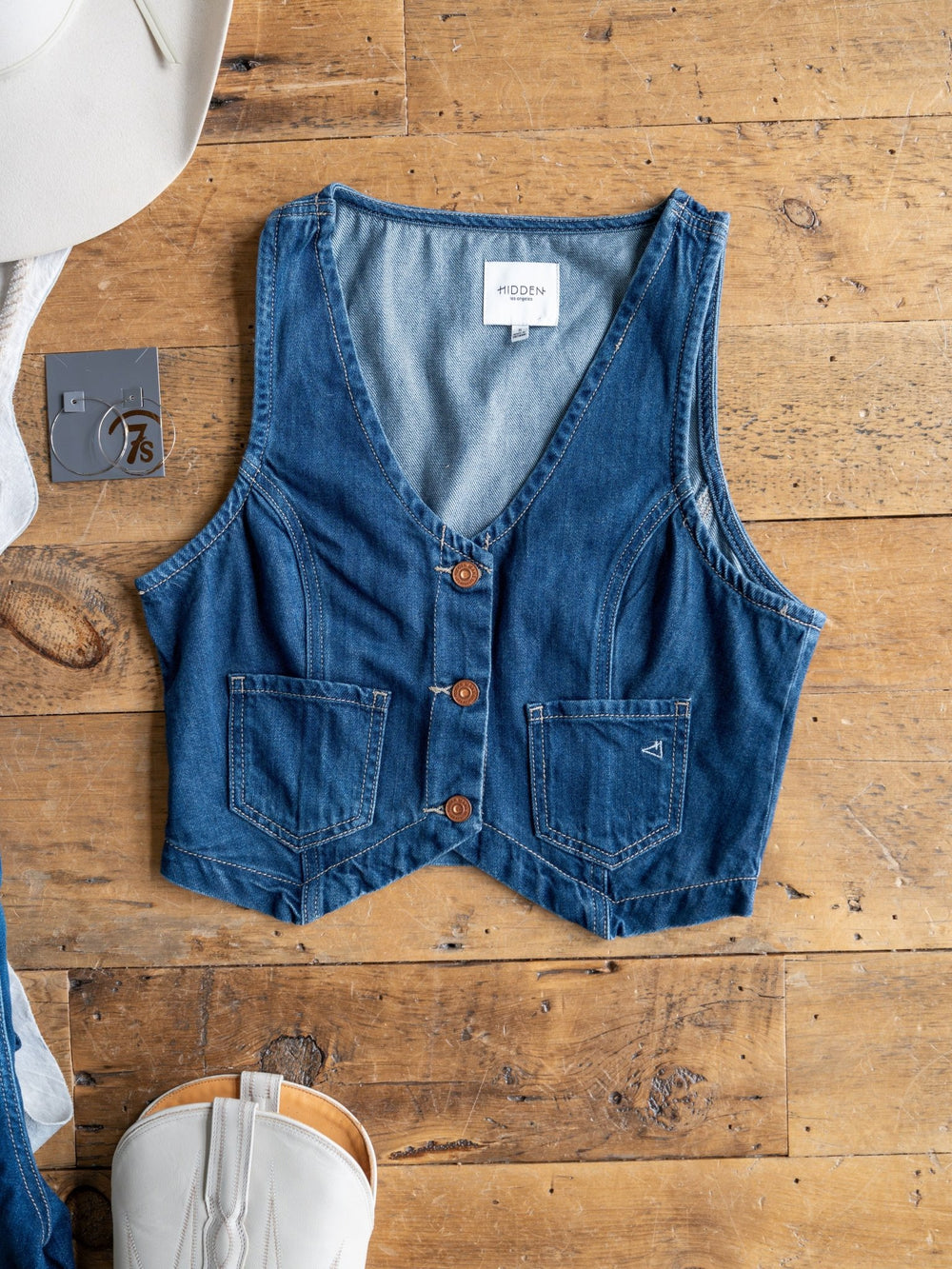 The Osage by Savannah Sevens western life{&}style, a sleeveless medium dark wash denim crop vest with three brown buttons and two front pockets, is displayed flat on wood beside a white cowboy hat, gray "7.5" tag, and white boot.