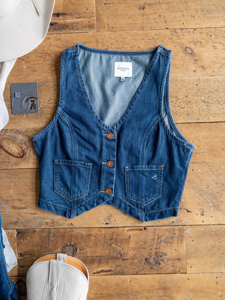 The Osage by Savannah Sevens western life{&}style, a sleeveless medium dark wash denim crop vest with three brown buttons and two front pockets, is displayed flat on wood beside a white cowboy hat, gray "7.5" tag, and white boot.