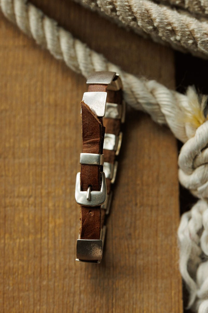A close-up of a brown leather watch strap with a silver buckle, placed vertically on wood, with thick coiled ropes and a Savannah Sevens western life{&}style Otoe Bracelet in the background.