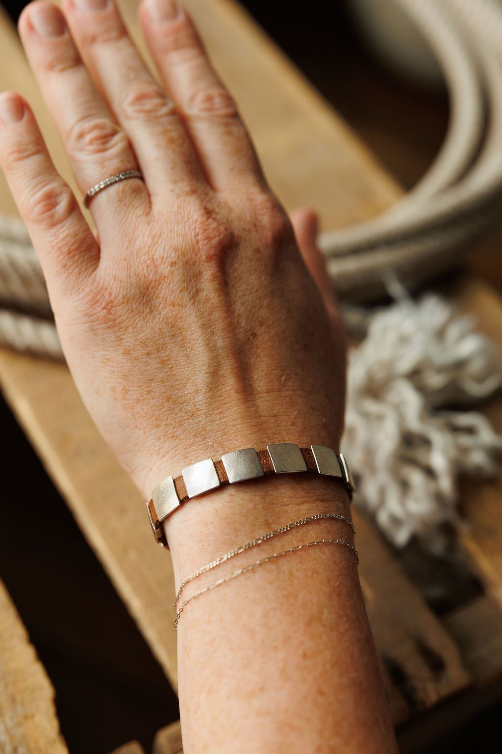 A hand wearing the Otoe Bracelet by Savannah Sevens western life{&}style, paired with two thin chain bracelets and a delicate ring, rests on a wooden surface with coiled rope in the background.