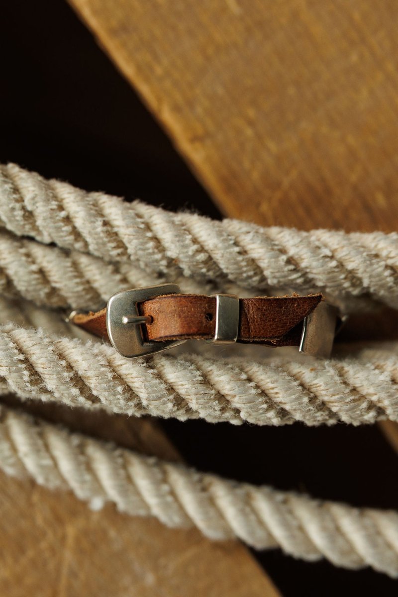 A close-up of the Otoe Bracelet by Savannah Sevens western life{&}style, a brown leather piece with a silver buckle, draped on thick white twisted rope atop wood, in Peyote Bird style.