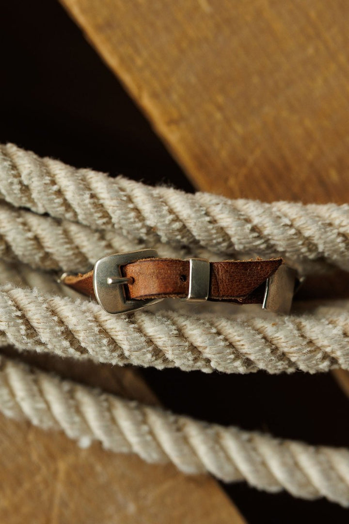 A close-up of the Otoe Bracelet by Savannah Sevens western life{&}style, a brown leather piece with a silver buckle, draped on thick white twisted rope atop wood, in Peyote Bird style.