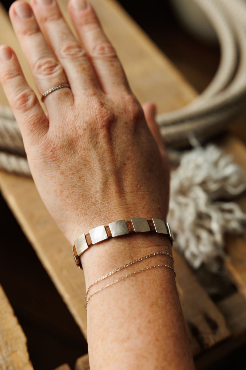 A hand wearing the Otoe Bracelet by Savannah Sevens western life{&}style, paired with two thin chain bracelets and a delicate ring, rests on a wooden surface with coiled rope in the background.