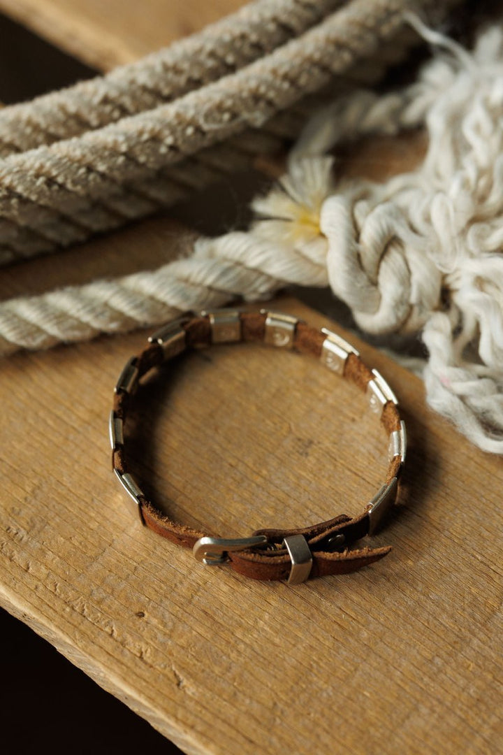 The Otoe Bracelet by Savannah Sevens western life{&}style, a brown leather accessory with metal accents, rests on a wooden surface next to coiled and braided ropes in natural light.