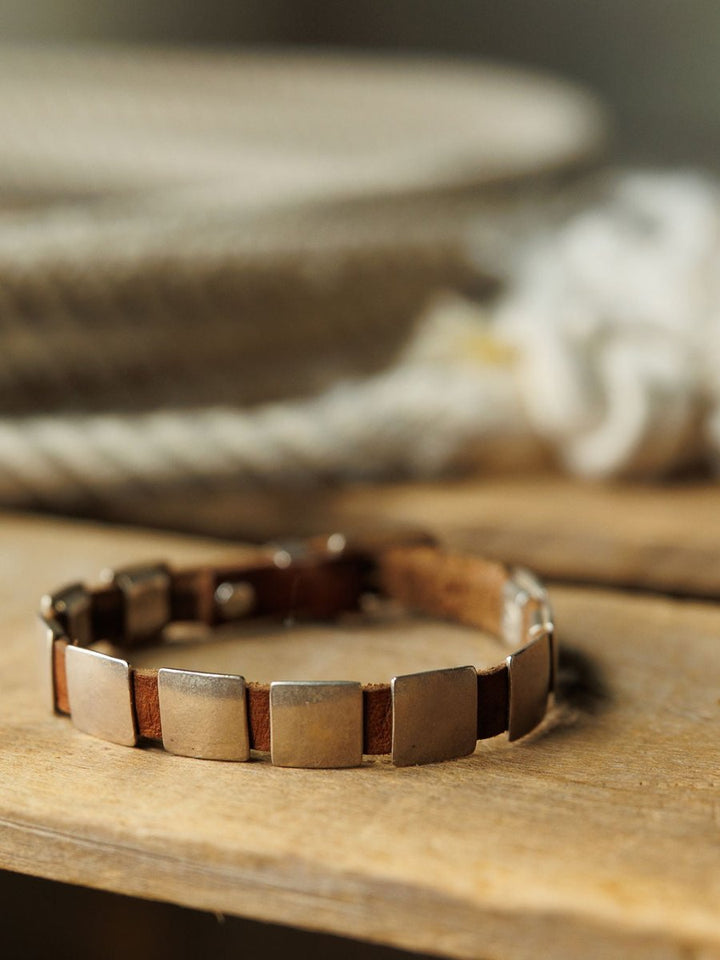 A close-up of the Savannah Sevens western life{&}style Otoe Bracelet, featuring leather and rectangular sterling silver plates, resting on wood with a blurred backdrop of coiled rope and soft neutral tones.
