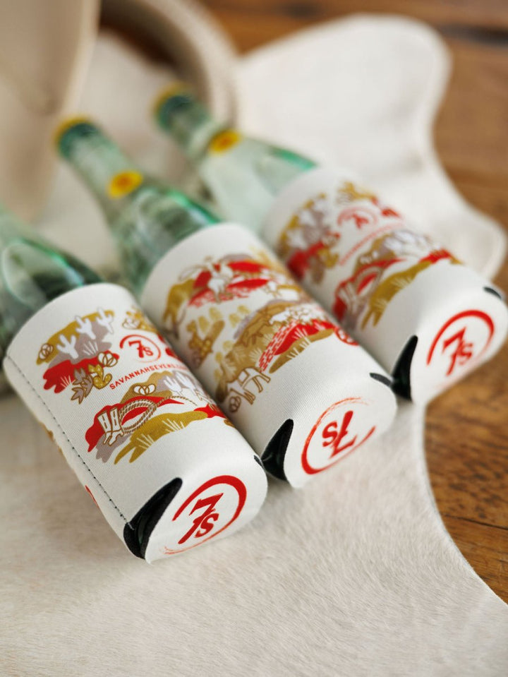 Three glass bottles with Savannah Sevens western life{&}style Out West Koozies, featuring artistic hand-drawn designs and a bold red 75, rest on a light surface with part of a hat in the background.