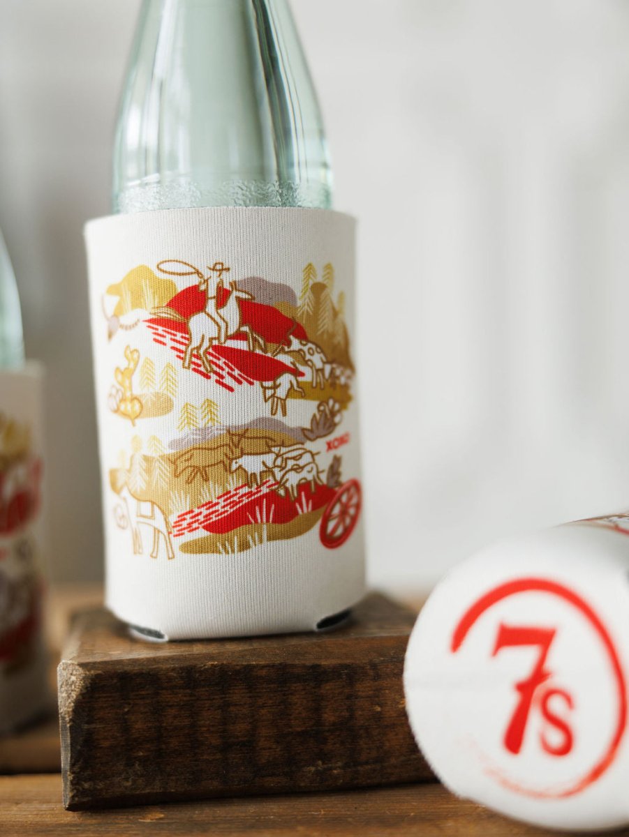 A glass bottle in the Out West Koozie by Savannah Sevens western life{&}style, featuring red and gold cowboy and horse designs, sits beside another artistic koozie with a bold red 7s logo. Both are displayed on a wooden surface.