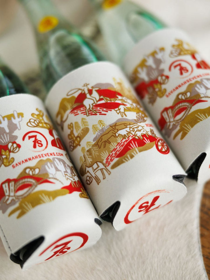 The Out West Koozie by Savannah Sevens western life{&}style includes three glass bottles in white and gold-patterned sleeves with hand-drawn horses, riders, wagons, “savannahsevens.com,” and a red “7S” logo on each sleeve.