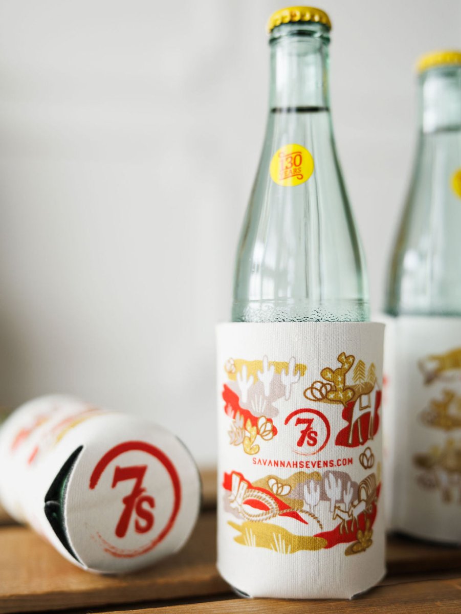 Two glass bottles with yellow caps, one upright and one lying down, are in Out West Koozies by Savannah Sevens western life{&}style. The white koozies have hand-drawn red and gold designs, the 7s logo, and savannahsevens.com printed on them.