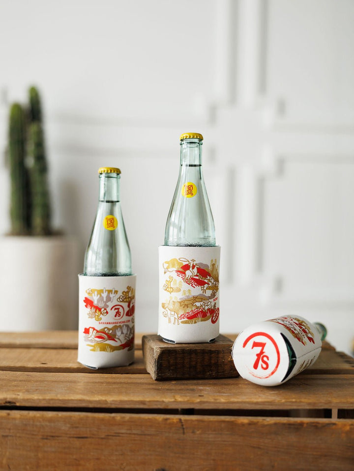 Three clear glass bottles with yellow caps are in white and red "Out West Koozie" holders by Savannah Sevens western life{&}style; two bottles stand upright, one is on its side. A potted cactus is blurred in the background.