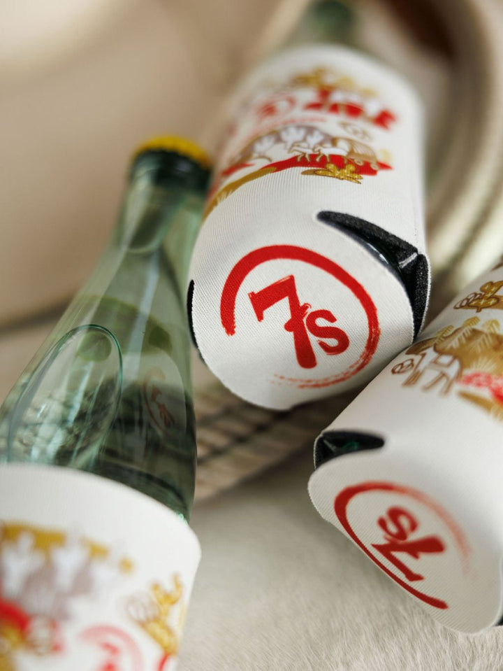 Close-up of glass soda bottles, some empty and some full, wrapped in Out West Koozie beverage holders by Savannah Sevens western life{&}style, featuring a red 7s logo and gold accents. A coiled rope appears in the background.