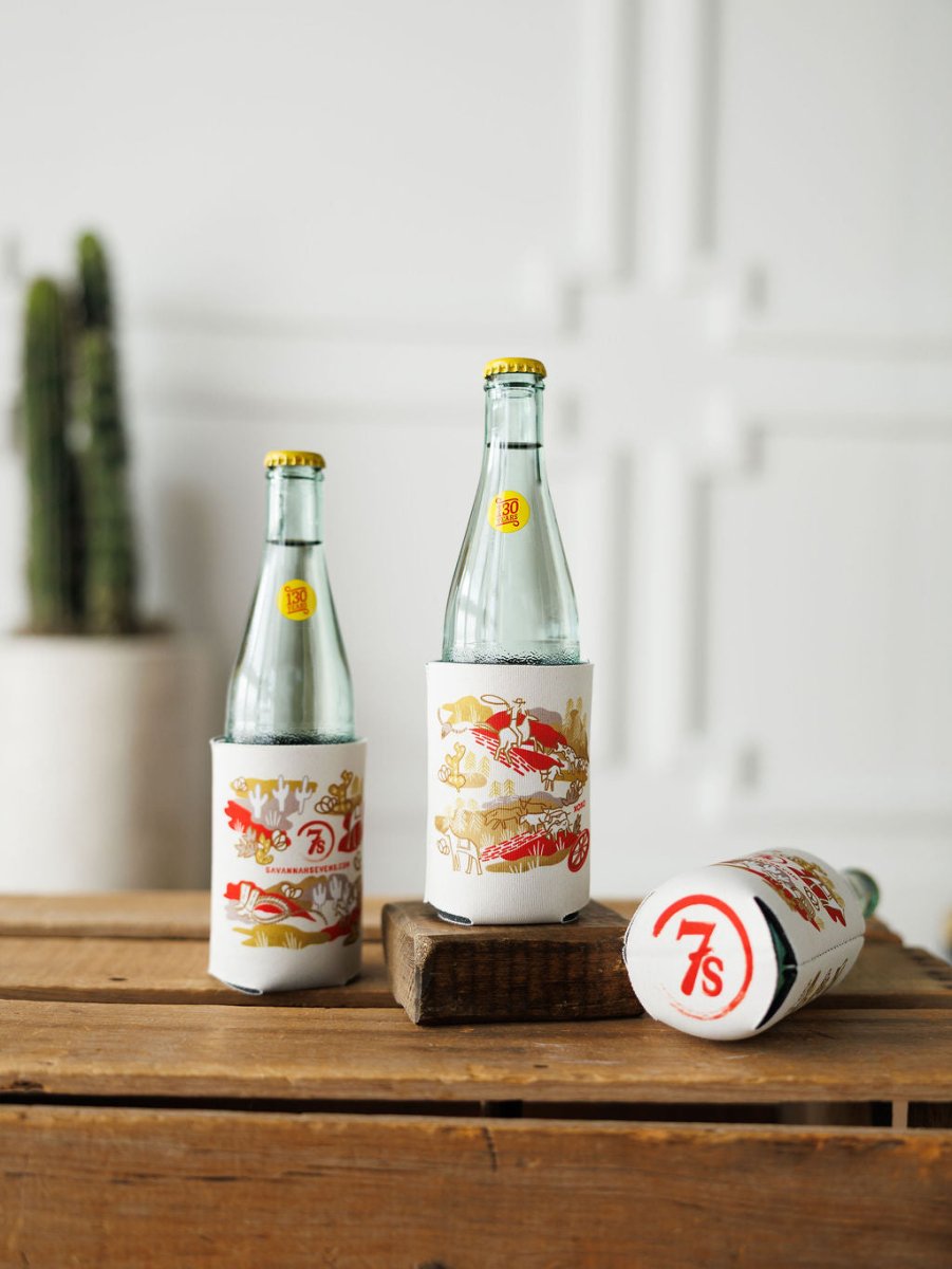 Three clear glass bottles with yellow caps are in white and red "Out West Koozie" holders by Savannah Sevens western life{&}style; two bottles stand upright, one is on its side. A potted cactus is blurred in the background.