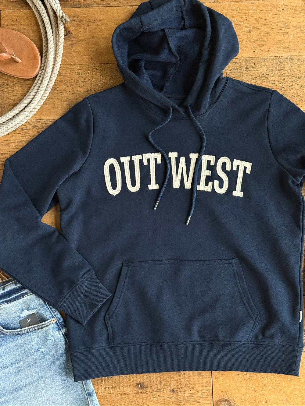 The Out West Midnight Navy Hoodie from Savannah Sevens western life{&}style is shown laid flat on a wooden floor alongside a rope, sandals, and light blue jeans. Featuring "OUT WEST" in bold white letters and designed with a relaxed fit, this hoodie is perfect for casual outings.
