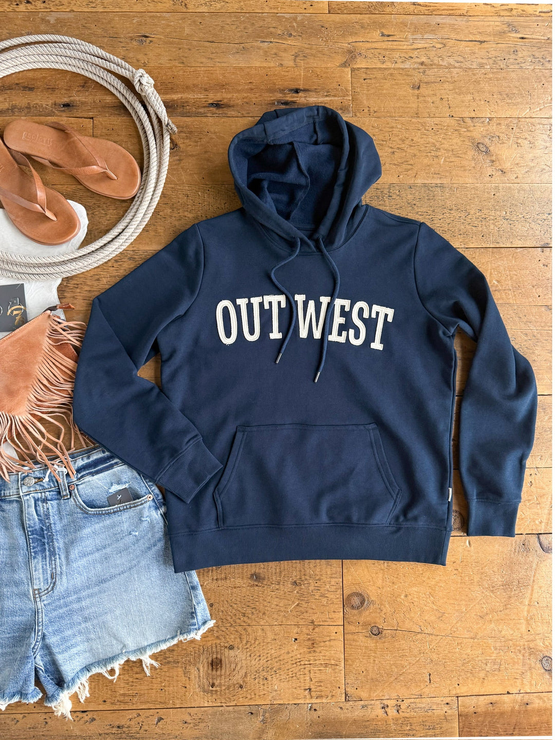 The Out West Midnight Navy Hoodie by Savannah Sevens western life{&}style, featuring "OUT WEST" in bold white lettering, is displayed on a wooden floor alongside cutoff denim shorts, a tan fringe bag, sandals, and a coiled rope.