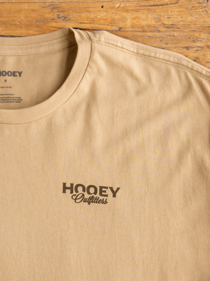 The Outfitters Mens Graphic Tee by Savannah Sevens western life{&}style is a tan men's graphic tee with "Hooey Outfitters" printed in black on the left chest, shown laid flat on a wooden surface.