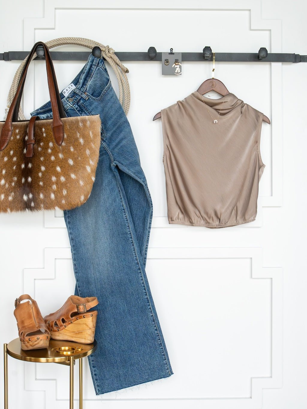 A pair of blue jeans, the Overland Cappaccino Satin Cowl Neck Top by Savannah Sevens western life{&}style, a brown spotted tote bag, and tan wedge sandals are neatly displayed against a white wall.