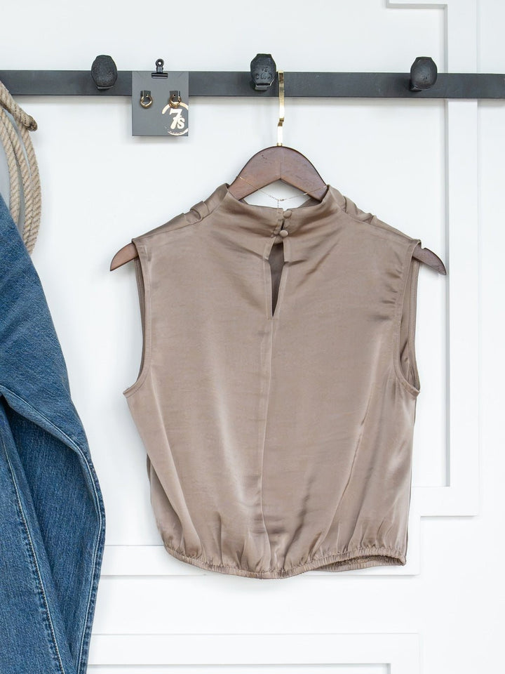 The Overland Cappuccino Satin Cowl Neck Top by Savannah Sevens western life{&}style is displayed sleeveless on a wooden hanger from a black wall hook, with blue jeans partially visible hanging to the left.