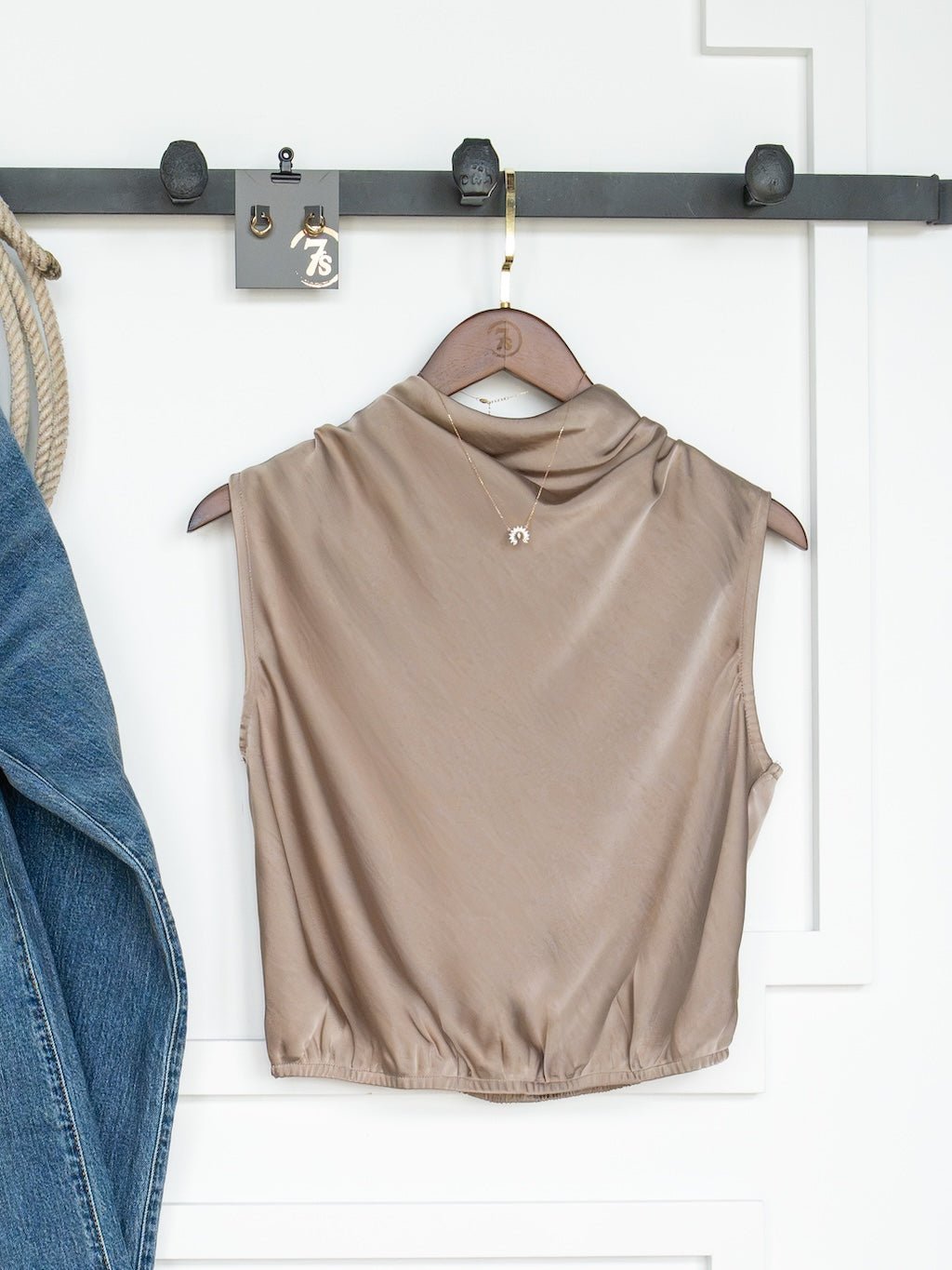 The Overland Cappaccino Satin Cowl Neck Top by Savannah Sevens western life{&}style hangs on a wooden hanger from a black hook rail. A gold necklace is draped over the top, and part of a blue denim jacket is visible on the left side of the image.