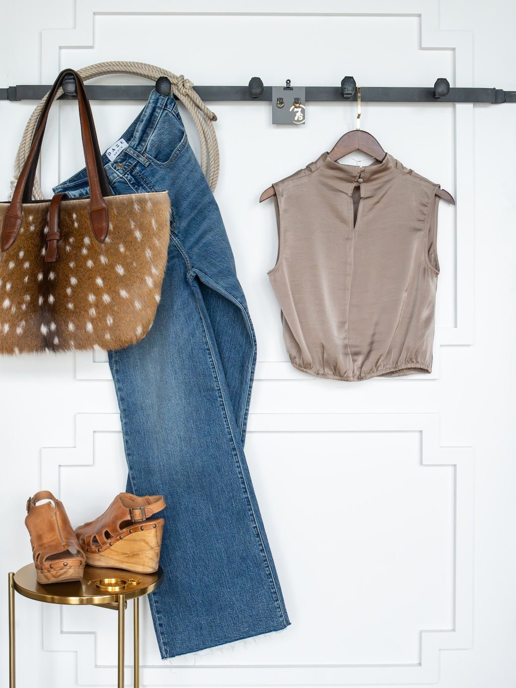 A sleeveless taupe blouse, blue jeans, brown wedge sandals, and a brown tote bag with white spots are stylishly displayed against a white paneled wall. The look is elevated with the chic touch of the Overland Cappaccino Satin Cowl Neck Top by Savannah Sevens western life{&}style.