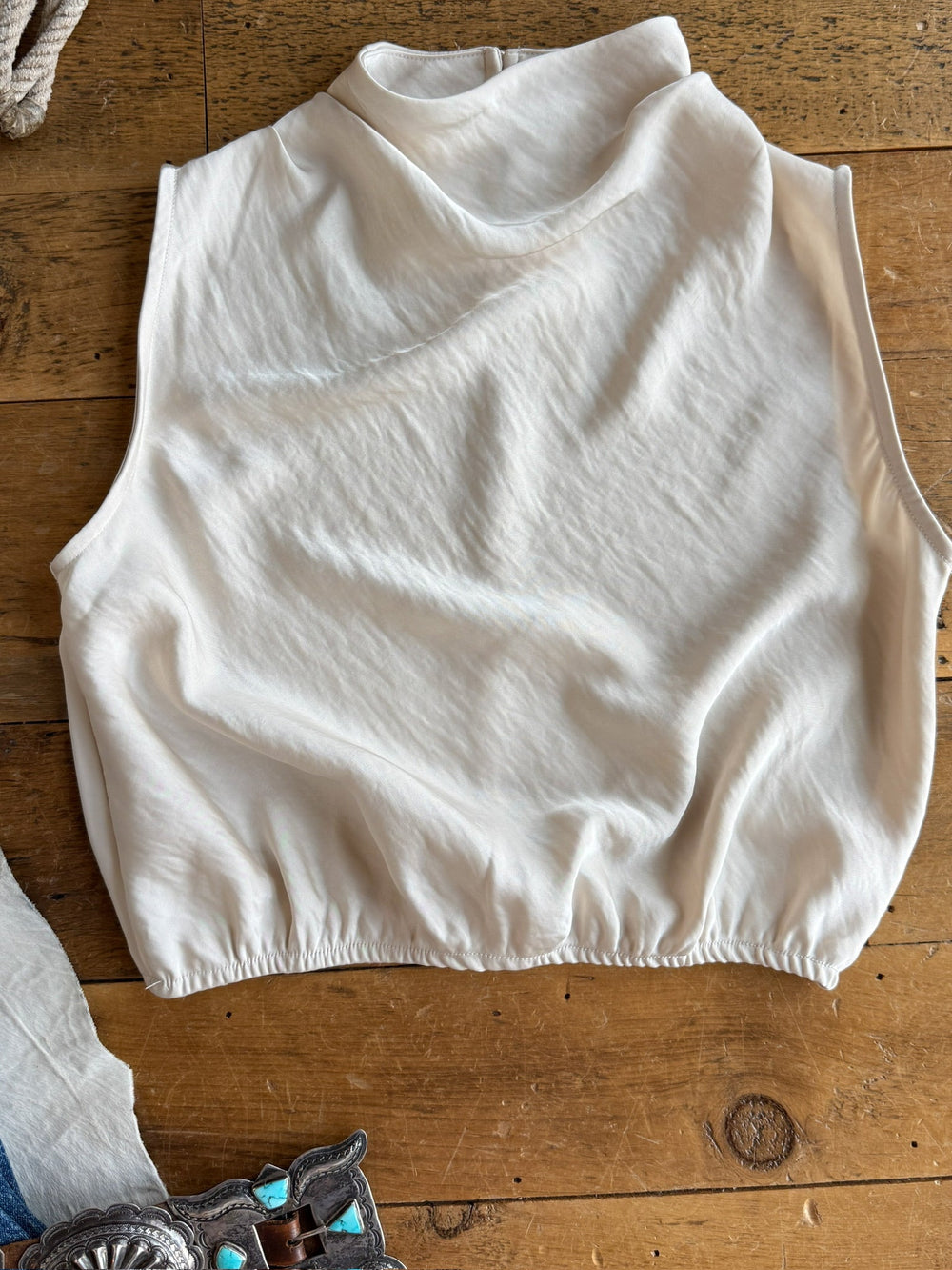 The Overland Champagne Satin Cowl Neck Top by Savannah Sevens western life{&}style, a sleeveless off-white crop top with a high neckline and elastic hem, is displayed flat on a wooden surface next to a belt adorned with turquoise stones.