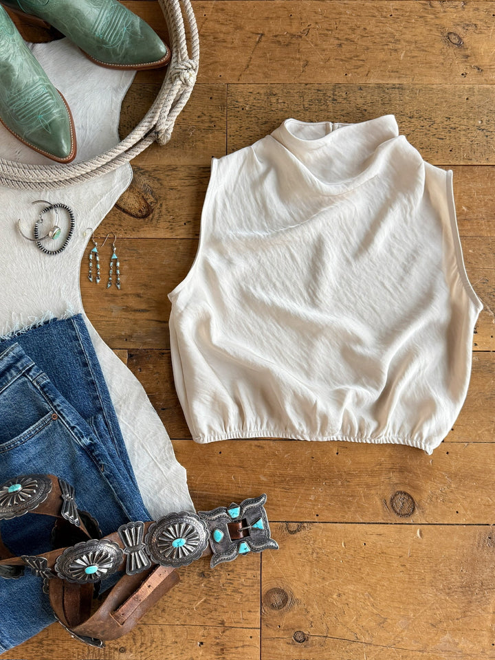 The Overland Champagne Satin Cowl Neck Top by Savannah Sevens western life{&}style is displayed on a wooden floor, artfully surrounded by blue jeans, a decorative belt, turquoise jewelry, green cowboy boots, and a coiled rope—Southwestern fashion accessories completing the stylish arrangement.