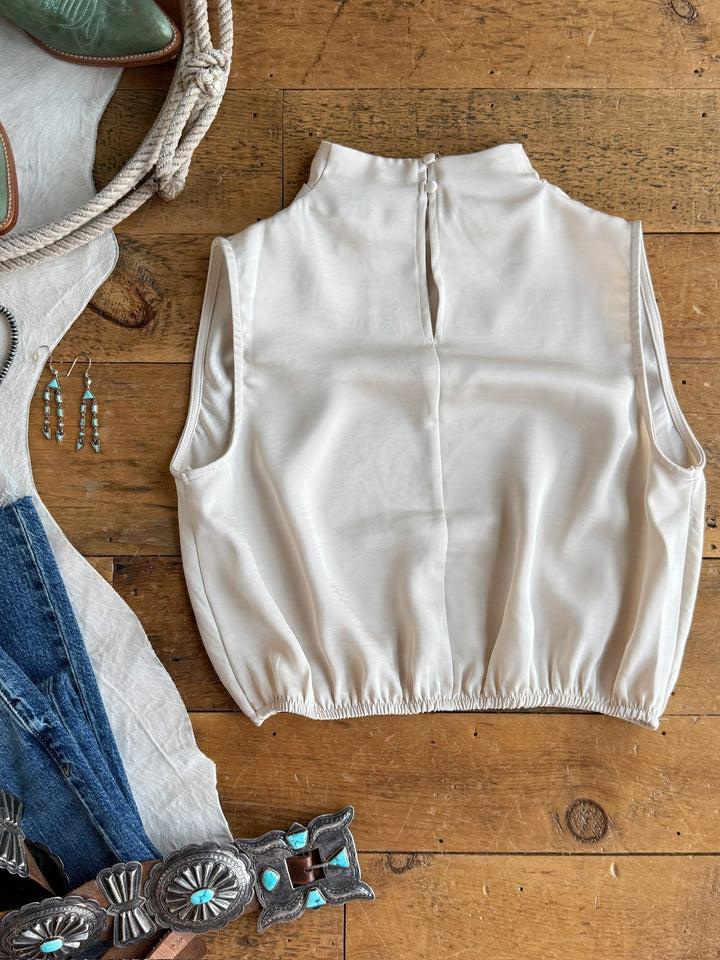 The Overland Champagne Satin Cowl Neck Top by Savannah Sevens western life{&}style rests on a wooden floor, accompanied by jeans, a turquoise concho belt, dangling earrings, boots, and a coiled rope.