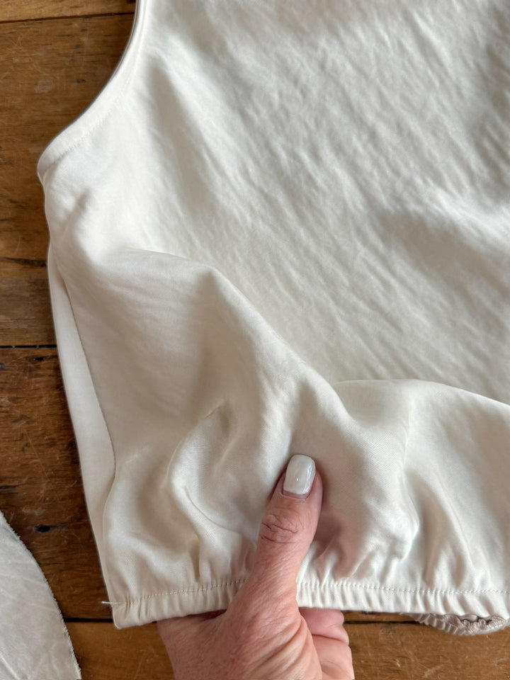 A hand holds the gathered elastic hem of the Overland Champagne Satin Cowl Neck Top by Savannah Sevens western life{&}style, displayed on a wooden surface.