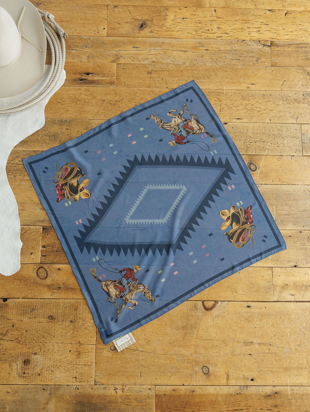 The Paducah Neck Scarf by Savannah Sevens western life{&}style, featuring a blue western print with cowboy hats and boots, is displayed on a wooden floor next to a white hat and white cloth.