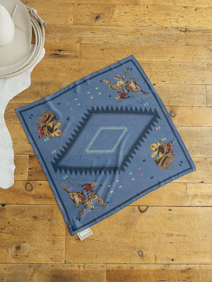 The Paducah Neck Scarf by Savannah Sevens western life{&}style, featuring a blue western print with cowboy hats and boots, is displayed on a wooden floor next to a white hat and white cloth.